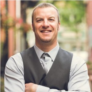 Photo of man, Jason Chaddock, smiling wearing a button up shirt and vest with arms crossed. Building and foliage in the background blurred.