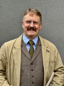 Photo of man with mustache and round glasses, Kurt Skinner, dressed as Teddy Roosevelt with blue button up shirt, green tie, brown vest, and over coat. Arms are down and hands out of frame presumably in his pockets.